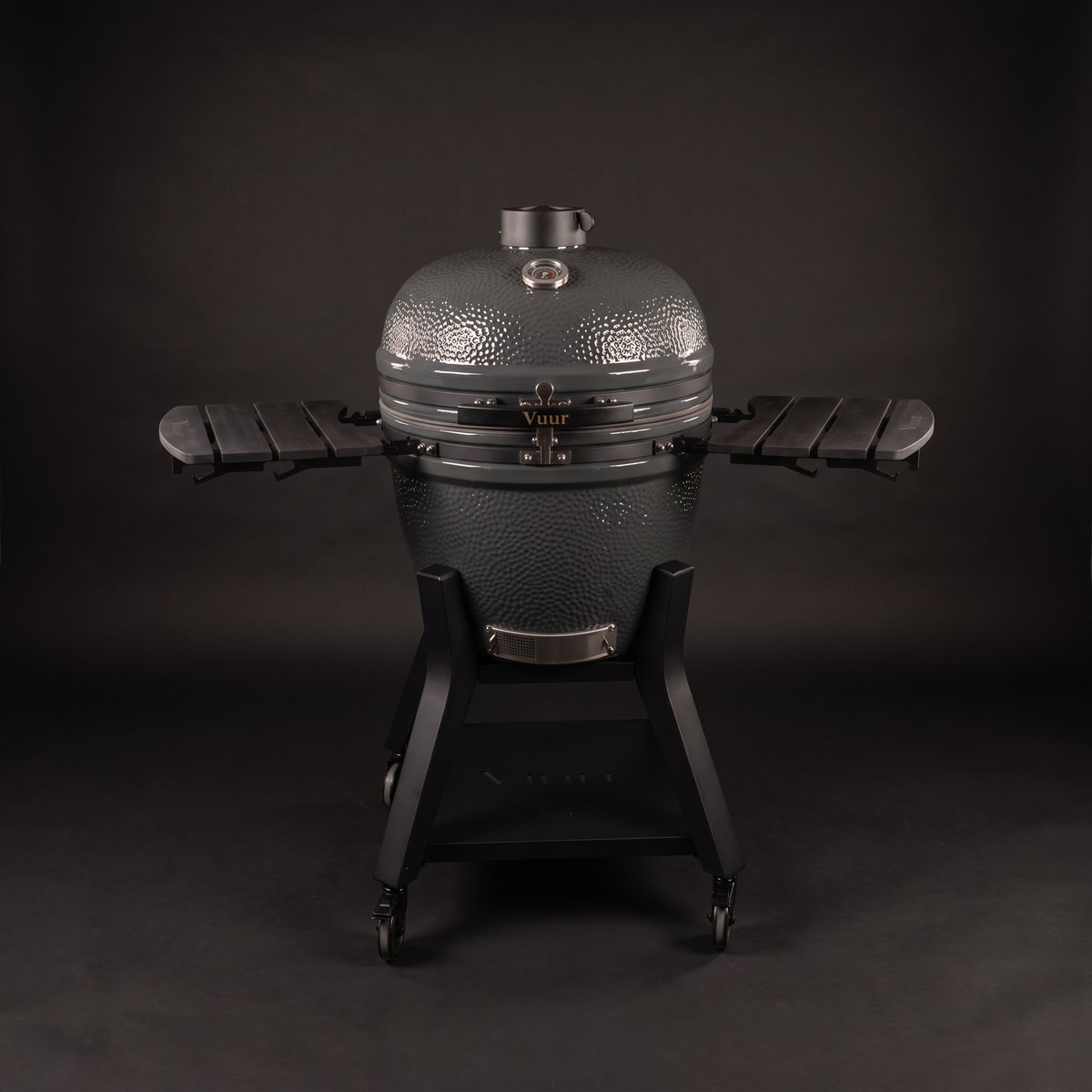 Grijze Vuur kamado 23 inch, gesloten, rechte voorkant, met de twee zwarte zijtafels uitgeklapt, op het stalen onderstel met wielen en de naam Vuur op de sluitband, op een donkere achtergrond.