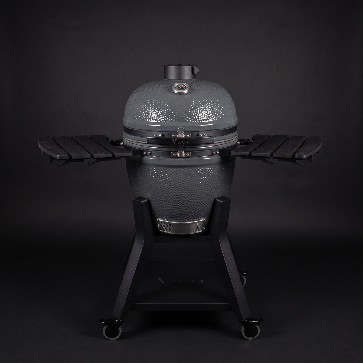 Grijze Vuur kamado 21 inch, gesloten, rechte voorkant, met de twee zwarte zijtafels ingeklapt en de naam Vuur op de sluitband, op een donkere achtergrond.
